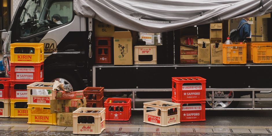 A black moving truck with its side open on a city street, loaded with cardboard boxes, plastic crates, and packing materials for a house relocation. The boxes include various sizes and are sealed with packing tape, with some displaying Japanese and English labels; others are plain or have printed logos of beverage brands such as Sapporo and Asahi. Several empty crates are stacked on the pavement in front of the van, some branded with Kirin and Sapporo. The interior of the truck shows additional boxes and packing supplies, indicating an ongoing loading process. A person wearing dark clothing is partially visible inside the truck, assisting with loading or organizing items. The scene includes natural daylight and wet pavement suggesting recent rain. This image illustrates furniture transport and packing activities typical of professional removals, supporting the services offered by Man with Van Farringdon in local home relocations and moving logistics.