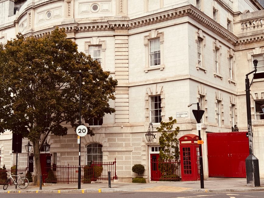 An exterior view of a historic white stone building with decorative architectural details and several tall, narrow windows. In front of the building, there is a sidewalk with a bicycle parked against a black iron fence, which encloses small landscaped areas with shrubs. A large leafy tree partially obscures the building's facade. On the street, there are two black street lamps, a traffic sign indicating a 20 mph speed limit, and a black trash bin. Adjacent to the building's entrance is a classic red British telephone box and a red metal gate or container near the corner. The scene is well-lit with natural daylight, suggesting a clear day. The image captures a typical urban environment suitable for home relocation, showing elements involved in furniture transport and packing and moving processes, with a focus on the building's exterior and street environment. The overall setting supports house removals services by highlighting the logistical aspects of loading and unloading in a city setting.