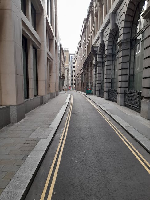 A narrow urban street with a curved asphalt road featuring double yellow lines along the center, flanked by stone-paved sidewalks on both sides. Tall, multi-storey buildings with classical architectural details, such as large arched windows and decorative stone facades, line the street. Some windows have metal railings, and the buildings are constructed from light-colored stone and darker, textured materials. The scene is well-lit with natural daylight, creating a neutral and professional atmosphere associated with house removals and moving logistics. The image illustrates a typical setting for residential or commercial property access, relevant to home relocation services offered by Man with Van Farringdon, showcasing street access for furniture transport and loading procedures before house removals.
