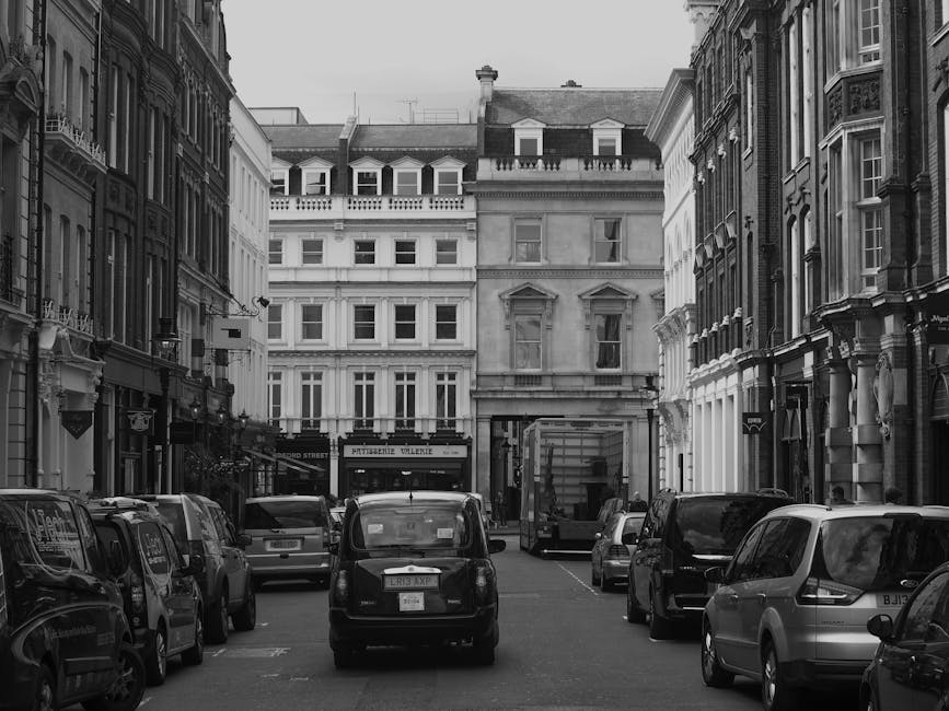 Black and white photograph of a narrow city street in Farringdon, showing parked cars on both sides and a delivery van in the center. The street is lined with historic multi-storey buildings featuring large sash windows, decorative stonework, and ornate facades. In the background, a taller building with a flat roof and several dormer windows is visible. The image captures the exterior environment where house removals and furniture transport may take place, highlighting the limited access typical for urban home relocation projects. Man with Van Farringdon occasionally performs moving services within this area, and the scene reflects the logistics involved in packing and loading household items onto vans for a house removal in a busy city centre setting.