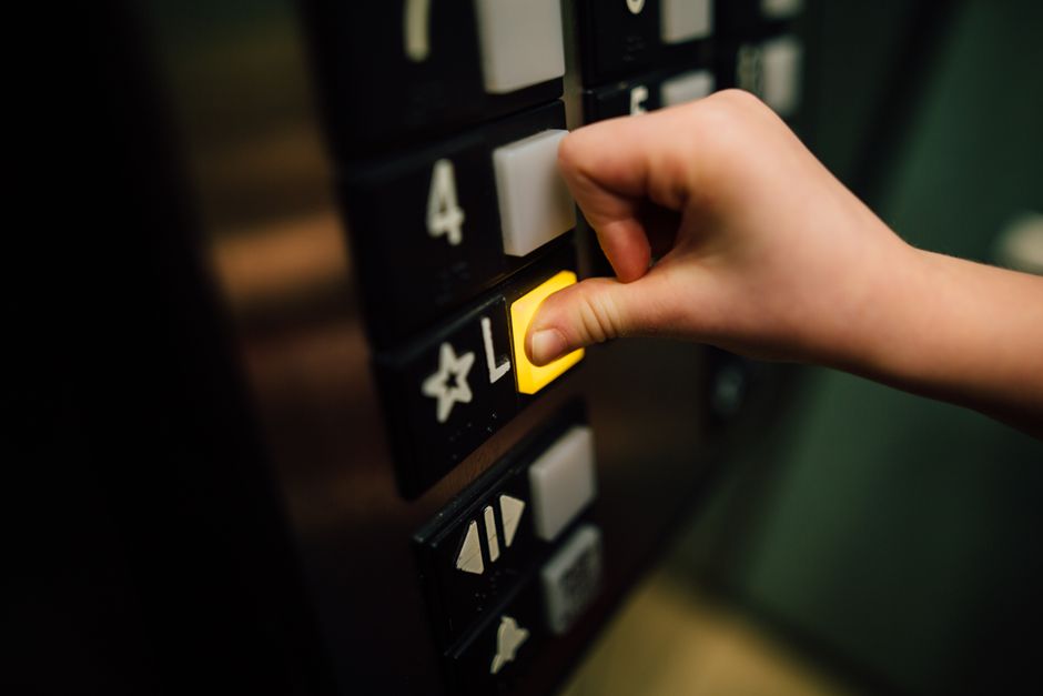 A close-up of a person's hand pressing a yellow button on an elevator control panel, which features black and white buttons numbered with digits and icons, situated inside a building corridor. The surrounding area includes other elevator buttons and indicator lights. The scene is part of a home relocation or moving process, where careful planning of elevator booking is important to prevent delays in the moving logistics. The ergonomic placement of the button and the lighting highlight the focus on access and operation of the elevator during the packing and furniture transport phases of a removals service. This visual detail is relevant to professional house removals, such as those provided by Man with Van Farringdon, emphasizing the importance of efficient building access during a house move.