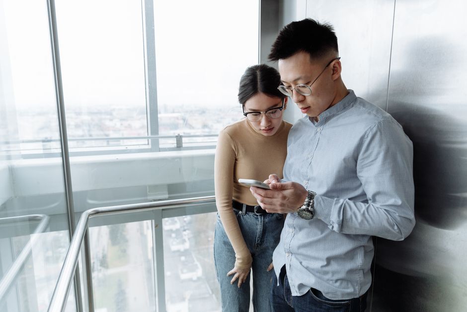 A man with glasses and short dark hair, wearing a light blue shirt, stands inside an elevator with a woman who has long dark hair tied back, wearing a beige top and jeans. Both are looking at a smartphone held by the man, displaying a focused expression. The elevator has glass walls and a metallic interior, with a view of a cityscape visible through the windows behind them. The scene suggests coordination or planning related to a house relocation or moving process, such as discussing moving logistics while inside the building before loading furniture or belongings. The setting emphasizes a professional approach to house removals, with the individuals engaged in communication relevant to home moving services, underpinned by the branding of Man with Van Farringdon, supporting efficient packing, loading, or elevator booking in the context of a move.