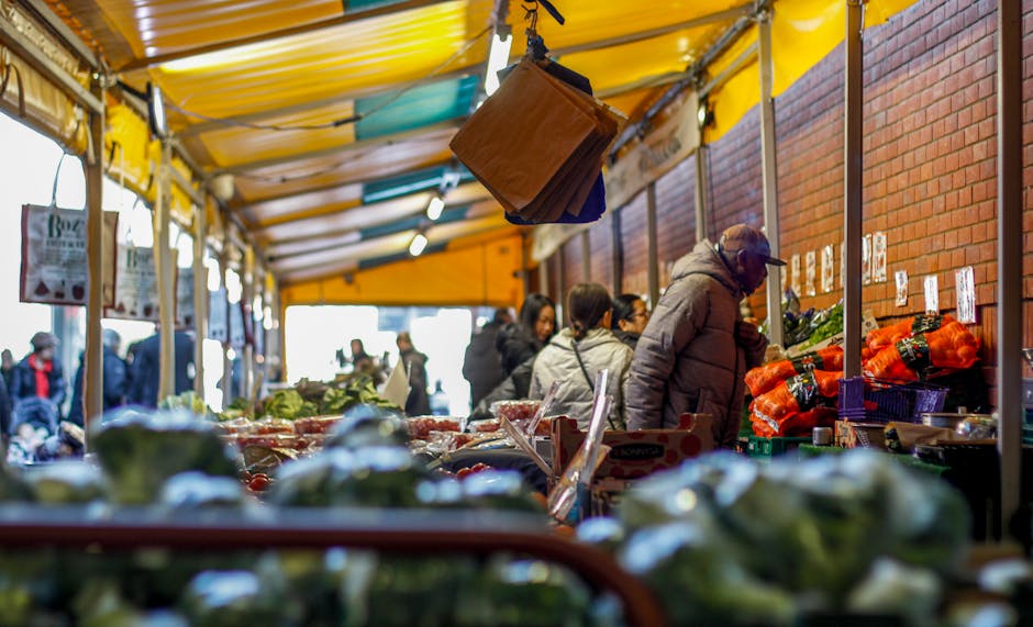 Inside a market stall with a yellow canopy, several people are browsing fresh vegetables and produce, including broccoli and carrots, displayed on wooden tables. The stall is situated against a red brick wall, and the processing area features cardboard boxes, plastic wrap, and packaging materials. Hanging from the ceiling of the stall is a brown wooden box or small cabinet, used for storage or display. In the background, some individuals are engaged in examining items, while others are walking past or shopping, with natural daylight illuminating the scene. The environment suggests a busy market setting, possibly during a market-day in Farringdon, and is relevant for a house or market move, with emphasis on the logistics of packing and transporting produce and belongings, as managed by a professional removals service such as Man with Van Farringdon.