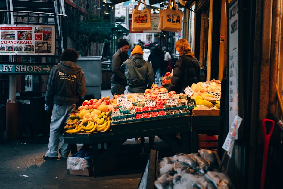 Inside a market stall with a yellow canopy, several people are browsing fresh vegetables and produce, including broccoli and carrots, displayed on wooden tables. The stall is situated against a red brick wall, and the processing area features cardboard boxes, plastic wrap, and packaging materials. Hanging from the ceiling of the stall is a brown wooden box or small cabinet, used for storage or display. In the background, some individuals are engaged in examining items, while others are walking past or shopping, with natural daylight illuminating the scene. The environment suggests a busy market setting, possibly during a market-day in Farringdon, and is relevant for a house or market move, with emphasis on the logistics of packing and transporting produce and belongings, as managed by a professional removals service such as Man with Van Farringdon.