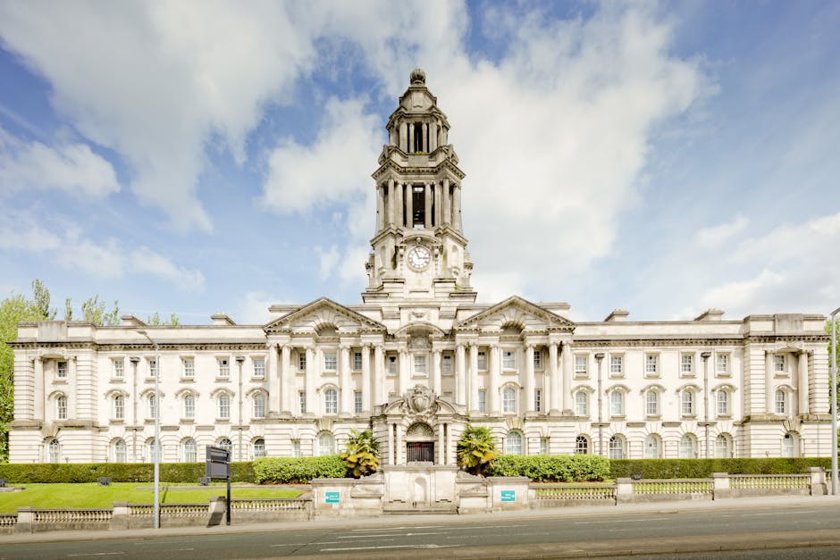 A large historic municipal building with a central clock tower, constructed from light-colored stone, featuring classical architectural details such as columns, decorative stonework, and arched windows. In the foreground, there is a paved street with a low metal fence, street signs, and green shrubbery along the building's base. The sky above is partly cloudy with blue patches, and the surrounding greenery includes trees with light green foliage. This setting reflects an urban environment suitable for house removals or home relocation services, with the building possibly serving administrative or civic functions. The image supports content related to property moving, packing, and furniture transport, as seen in the presence of a well-maintained exterior and prominent architectural features, aligned with the context of securing permits for removals in Farringdon, as referenced in the page title for Man with Van Farringdon.