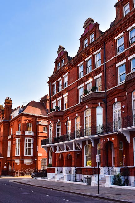 A row of red-brick residential buildings along a quiet street, featuring large windows and decorative balconies with black wrought iron railings. The buildings are several stories high, with the upper floors showing dormer windows and ornate architectural details. The street in front is paved, with parked cars and a pedestrian sidewalk, and a clear blue sky overhead indicates daytime. In the foreground, there is no visible furniture or moving equipment, but the setting suggests an area where house removals or home relocation services, like those offered by Man with Van Farringdon, could take place.
