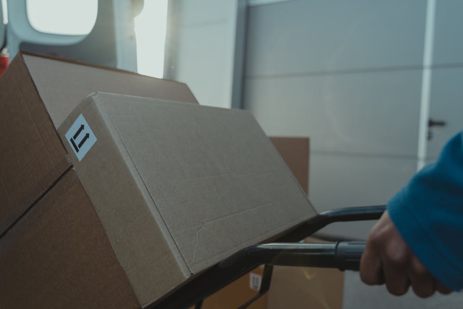 A person wearing a blue long-sleeve shirt is pushing a black hand truck loaded with three cardboard boxes, which are stacked one on top of the other with visible packing tape. The boxes are plain brown, and one has a small white label. They are inside a well-lit room with natural light coming through large windows or glass doors in the background, and a white door or wall is visible behind. The person appears to be in the process of loading or transporting the boxes, likely as part of home relocation or furniture transport arranged by Man with Van Farringdon. The scene depicts a typical packing and loading activity during house removals, with the boxes secured for safe transport, potentially heading toward a van parked outside, as part of a professional removal process focused on efficient packing and moving logistics.