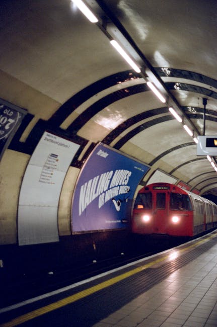 Inside a London underground station tunnel, a red train is arriving or departing, with its headlights on. The curved ceiling features dark lines and white lighting panels, and there are various posters and advertisements on the underground walls, including a prominent blue one displaying the words 'MAKE MOVES.' The platform is visible in the foreground, with tactile paving strips along the edge for safety. This setting indicates a busy transit environment typical of urban house removals and furniture transport within London, where efficient loading and unloading of moving vehicles, such as those used by Man with Van Farringdon, are common during home relocation processes.