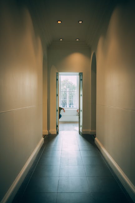A narrow, well-lit interior corridor with smooth beige walls and a dark grey tiled floor, leading to a blue door at the end that opens to a bright balcony with outside greenery visible through large glass windows. The ceiling features three recessed spotlights evenly spaced, illuminating the hallway. At the threshold of the door, there is a person partially visible, wearing dark clothing and handling a cardboard box or packing material, suggesting a home relocation or moving process. The scene aligns with furniture transport and packing activities typical in house removals, with the narrow space presenting challenges for moving large items through confined doorways, as managed by companies like Man with Van Farringdon.