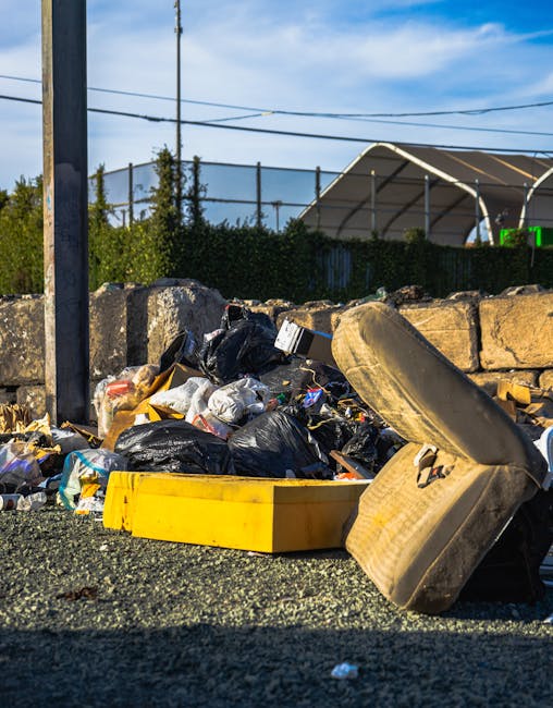 An outdoor scene displaying a large pile of mixed waste materials including black plastic rubbish bags, cardboard boxes, and discarded furniture such as an old, stained armchair with fabric upholstery and a broken, partially overturned yellow plastic container. The waste is situated on a gravel surface against a stone wall, with a metal pole and overhead electrical wires visible in the foreground. In the background, there is a hedge, a partially enclosed sports facility with a curved roof, and a clear blue sky with some wispy clouds, indicating daytime. The image illustrates improper waste disposal which relates to legal considerations around waste management and removal services, as provided by Man with Van Farringdon, specializing in house removals and furniture transport, including packing and loading processes within property relocation contexts.