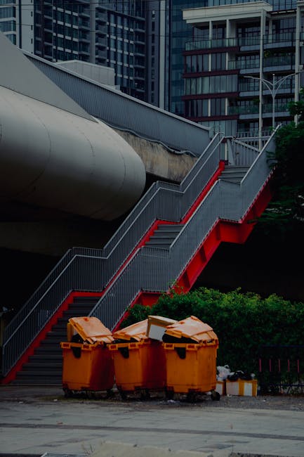 The image depicts an outdoor urban setting with a large, metallic, cylindrical pipeline elevated above ground, supported by structural elements and adjacent to modern multi-storey buildings with glass facades. A set of outdoor metal stairs with red-painted sides and silver handrails runs along the right side, leading from the ground level up to the elevated pathway or platform. At the base of the stairs, several large yellow and orange skip bins are positioned on a paved surface, filled with various waste materials and packaging debris, including cardboard boxes, plastic wrapping, and other refuse, indicating a waste disposal or clearance activity. Behind the bins, there are green shrubs and small trees, providing some natural contrast to the industrial and residential environment. The scene appears to be during daylight with natural lighting, and the setting suggests a typical urban area where house removals or building clearance services, possibly provided by Man with Van Farringdon, are involved in packing and loading processes related to property relocations or waste disposal near Farringdon.