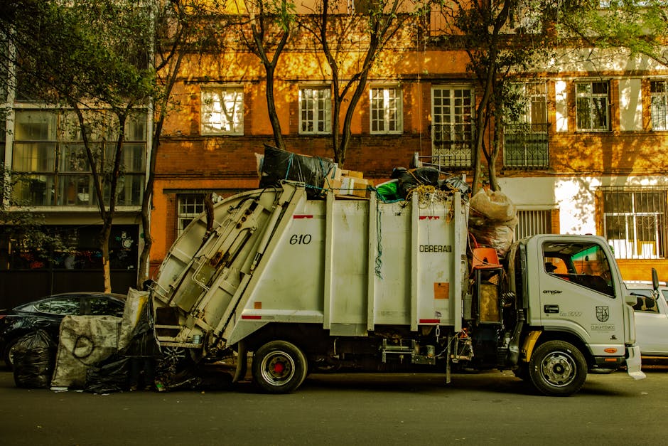 An outdoor scene displaying a large pile of mixed waste materials including black plastic rubbish bags, cardboard boxes, and discarded furniture such as an old, stained armchair with fabric upholstery and a broken, partially overturned yellow plastic container. The waste is situated on a gravel surface against a stone wall, with a metal pole and overhead electrical wires visible in the foreground. In the background, there is a hedge, a partially enclosed sports facility with a curved roof, and a clear blue sky with some wispy clouds, indicating daytime. The image illustrates improper waste disposal which relates to legal considerations around waste management and removal services, as provided by Man with Van Farringdon, specializing in house removals and furniture transport, including packing and loading processes within property relocation contexts.