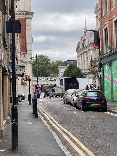 A narrow urban street scene during daytime with parked cars along the pavement, including a black vehicle closest to the camera and several other cars ahead. The street is lined with historic-style bu