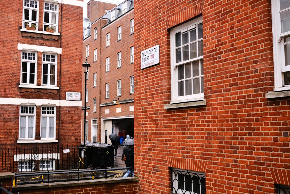 A narrow urban street scene during daytime with parked cars along the pavement, including a black vehicle closest to the camera and several other cars ahead. The street is lined with historic-style bu