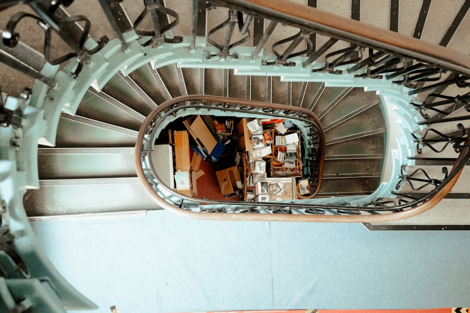 A spiral staircase with black metal railings and wooden handrails, viewed from above looking down into the stairwell. The staircase curves around a central void filled with various cardboard boxes, sm